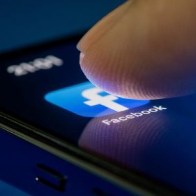 A fingertip pushes on the Facebook App on a mobile phone screen.
